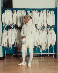 Fencer in white uniform and protective mask standing in front of clothing racks filled with white gear by Braylen Dion