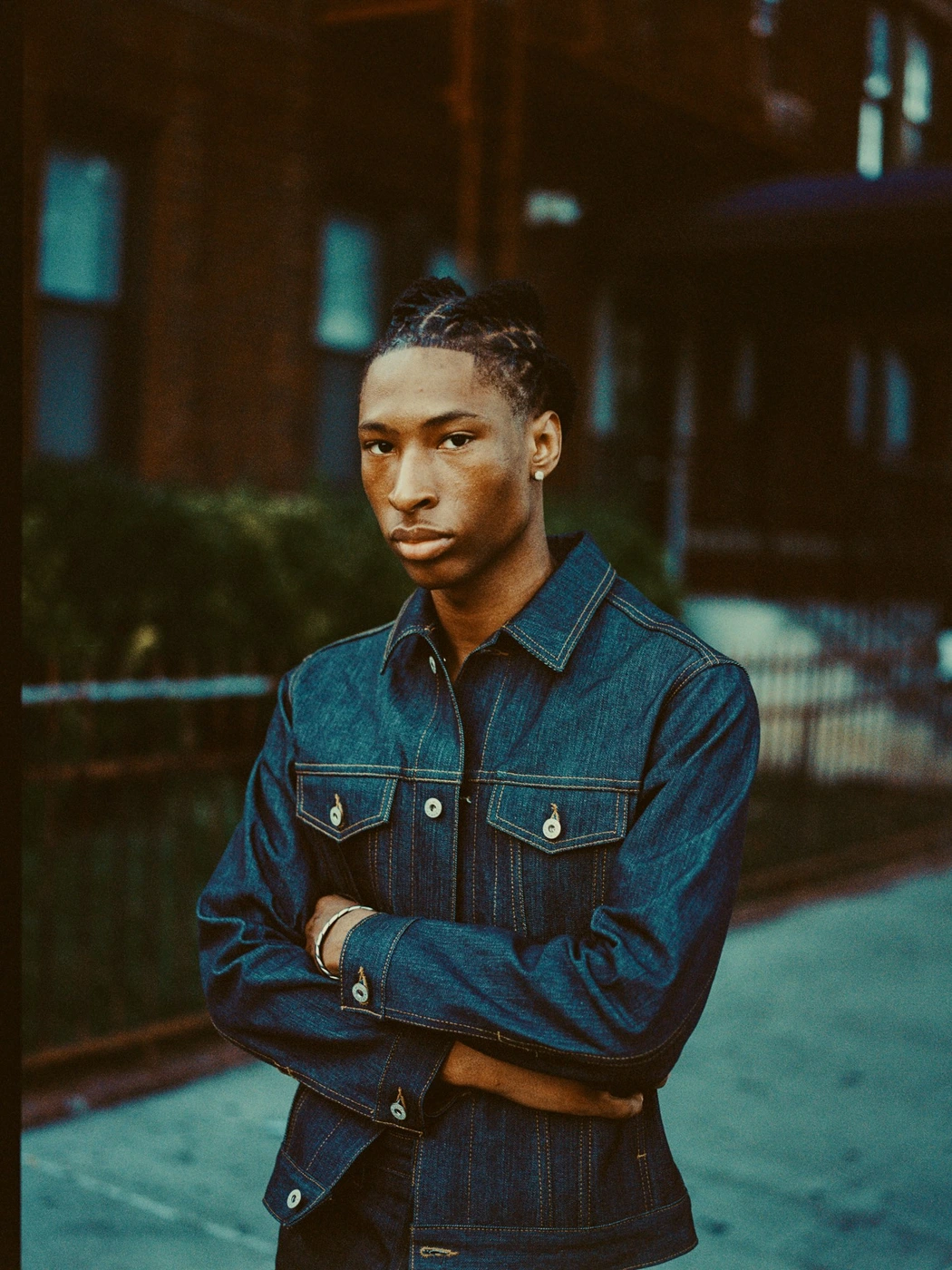 Black man wearing a dark denim jacket with arms crossed outdoors in front of a building facade.