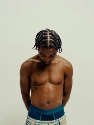 Shirtless man with braided hair looking down against a neutral studio background, photographed by Braylen Dion.
