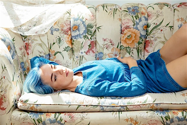 A young woman with blue hair lies on a floral sofa, wearing a denim dress, in a softly lit indoor setting with a relaxed pose.