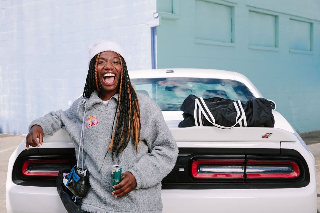 A smiling Black woman with braided hair stands in front of a white car, holding a Red Bull energy drink can, outdoors in daylight.