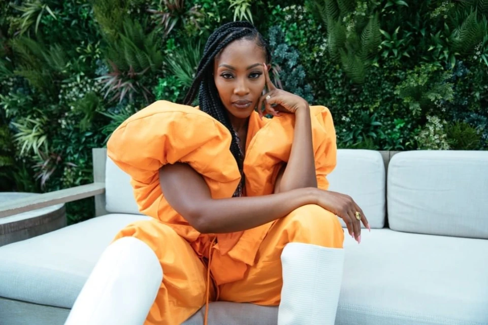 A stylish black woman with braided hair sits on a white couch in a bold orange jumpsuit, paired with white boots, against a leafy green wall.