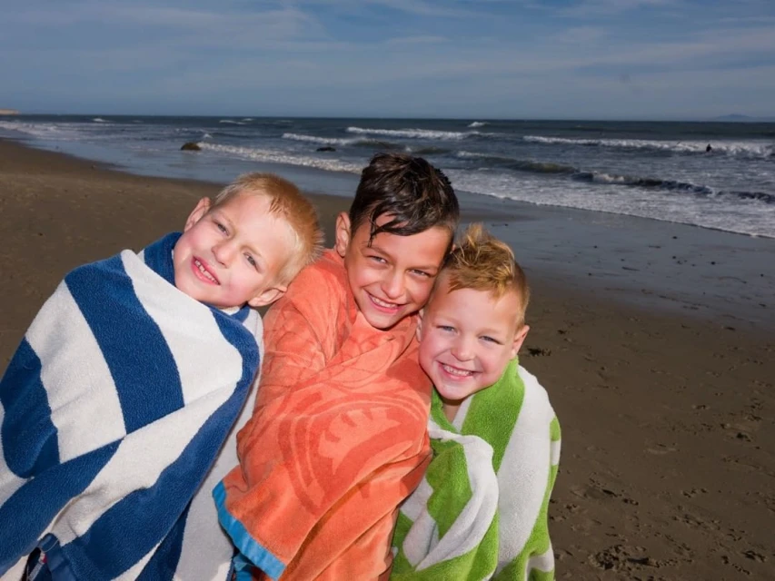 Three smiling children wrapped in colorful towels on a sandy beach with gentle waves, creating a joyful seaside portrait.