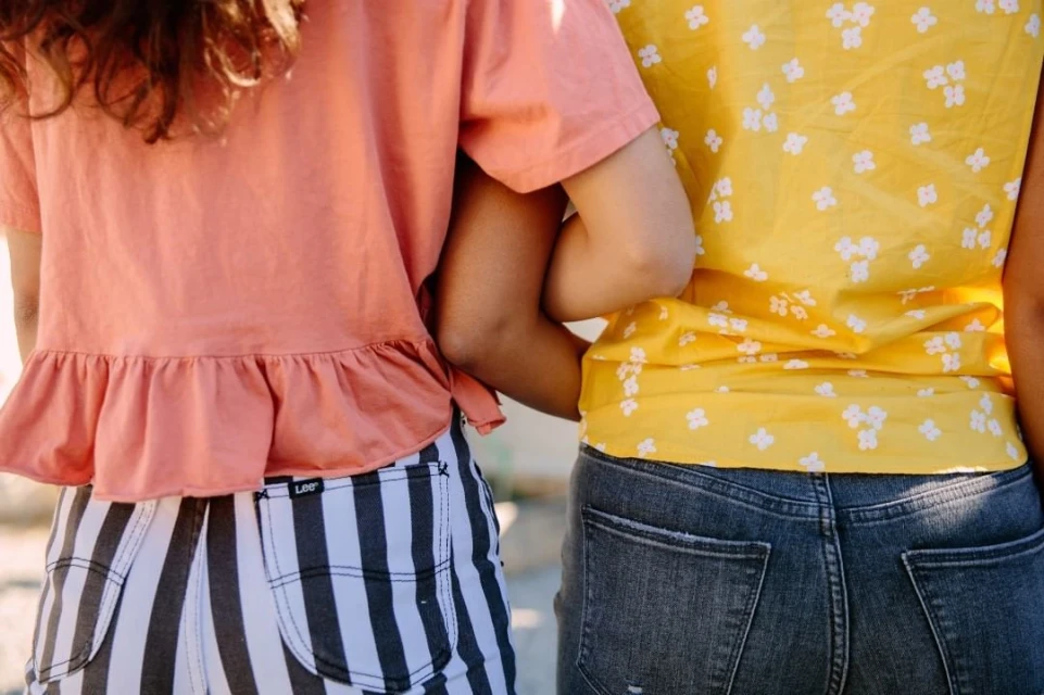 Two women stand close together outside in a sunny outdoor setting. One wears a peach top and striped pants, the other yellow floral top and jeans.