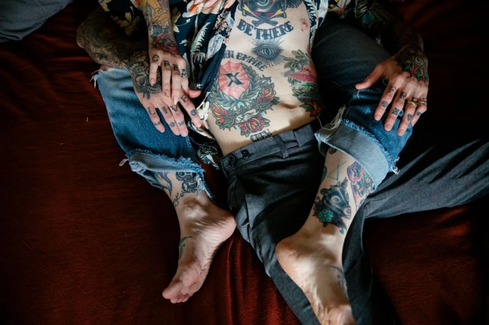 Tattooed person lies on red fabric with bare torso, hands, feet visible, wearing floral shirt and jeans.