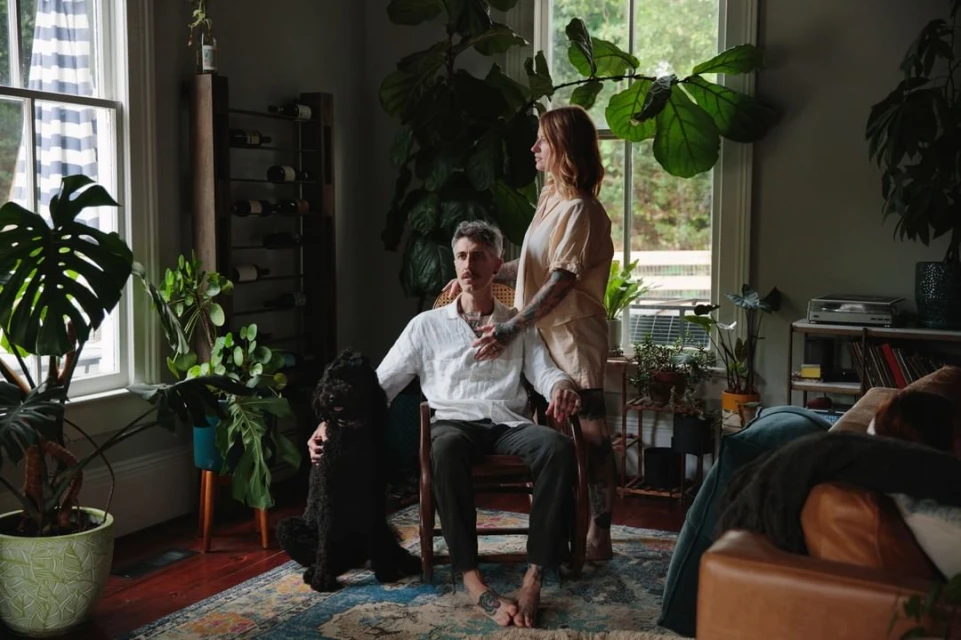 Indoor portrait of a tattooed couple and their black dog sitting on a rug surrounded by potted plants near windows and furniture.