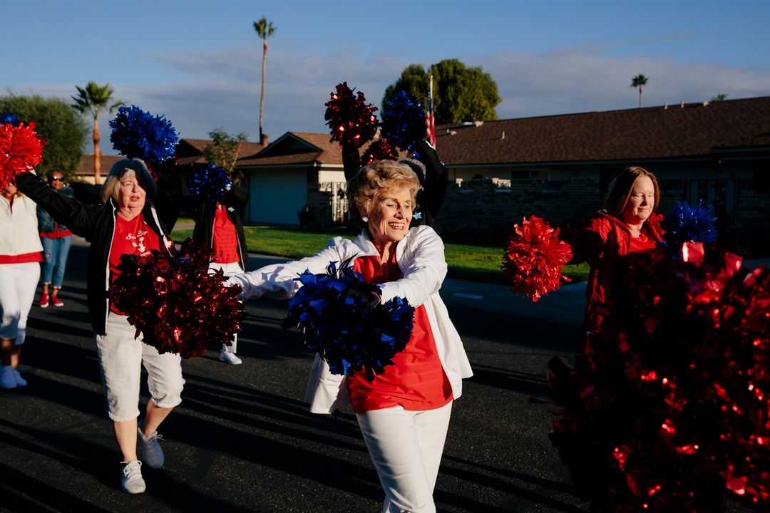 Group of mature women in matching white pants and colorful tops march on a suburban street holding red and blue pom-poms in bright sunlight.