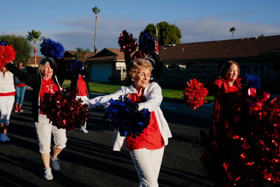 Group of mature women in matching white pants and colorful tops march on a suburban street holding red and blue pom-poms in bright sunlight.
