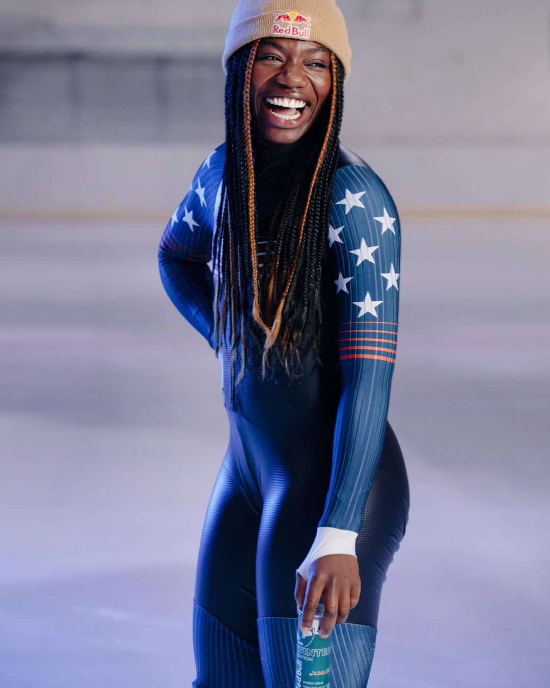 A smiling African American woman in a navy blue speed skating suit with white stars and a tan beanie poses indoors, holding a beverage.