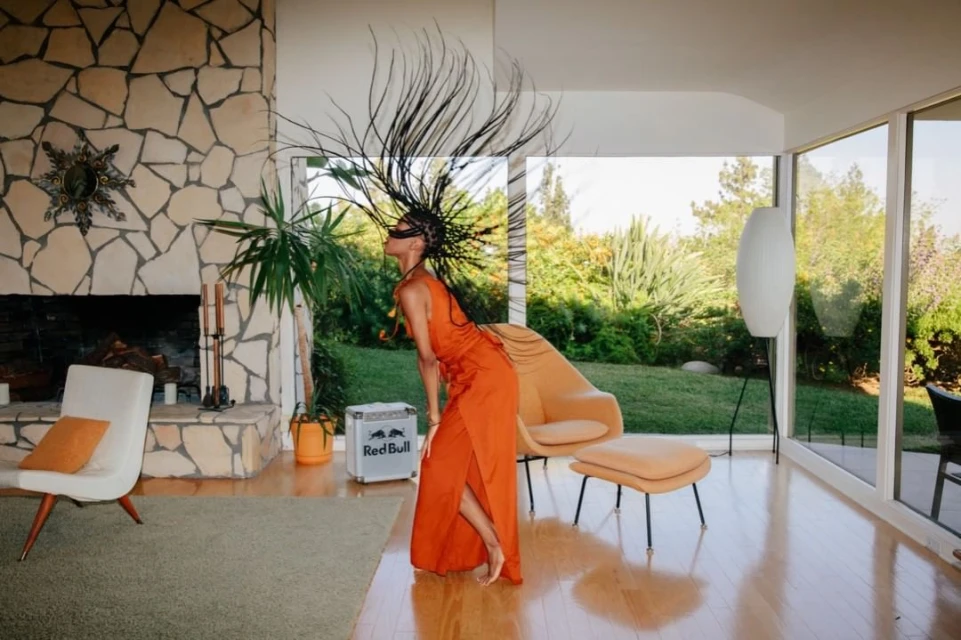 Woman in orange dress with elaborate hairstyle poses in a modern living room with stone fireplace and view of a lush green yard.