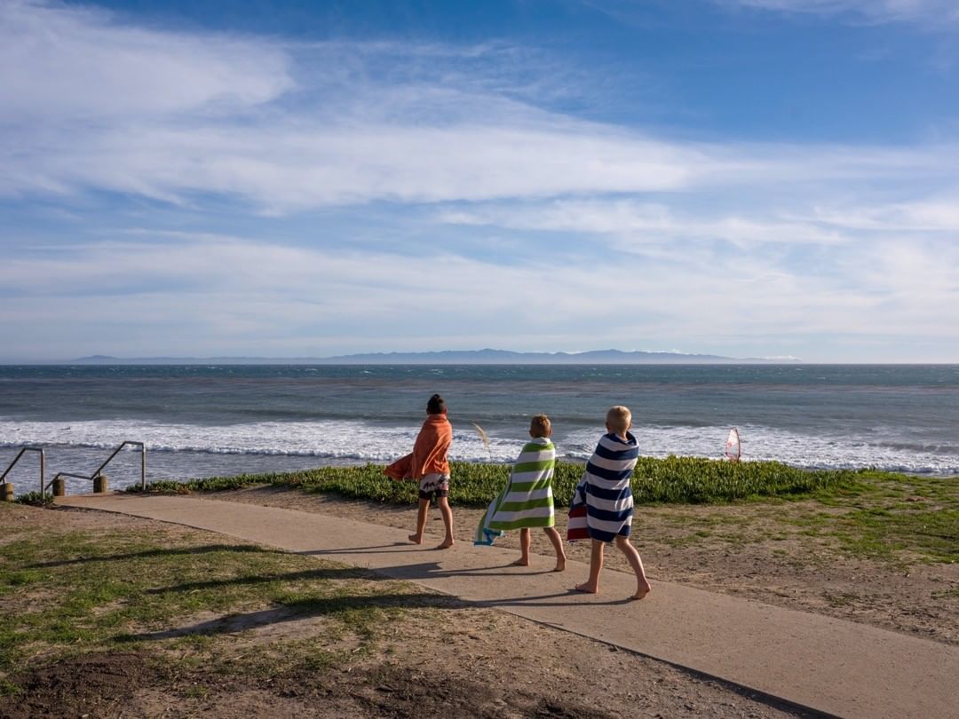 Three young figures, wrapped in towels, walk along a sandy path toward the ocean under a partly cloudy sky on a sunny day.