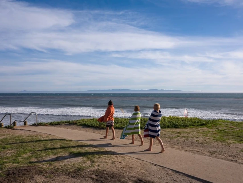 Three young figures, wrapped in towels, walk along a sandy path toward the ocean under a partly cloudy sky on a sunny day.