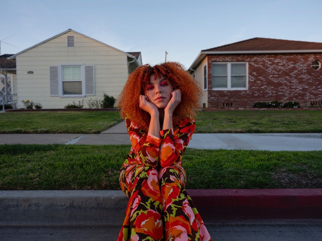 Red-haired woman in a floral dress sits on a curb in front of suburban houses on a sunny day, captured in a medium outdoor shot.