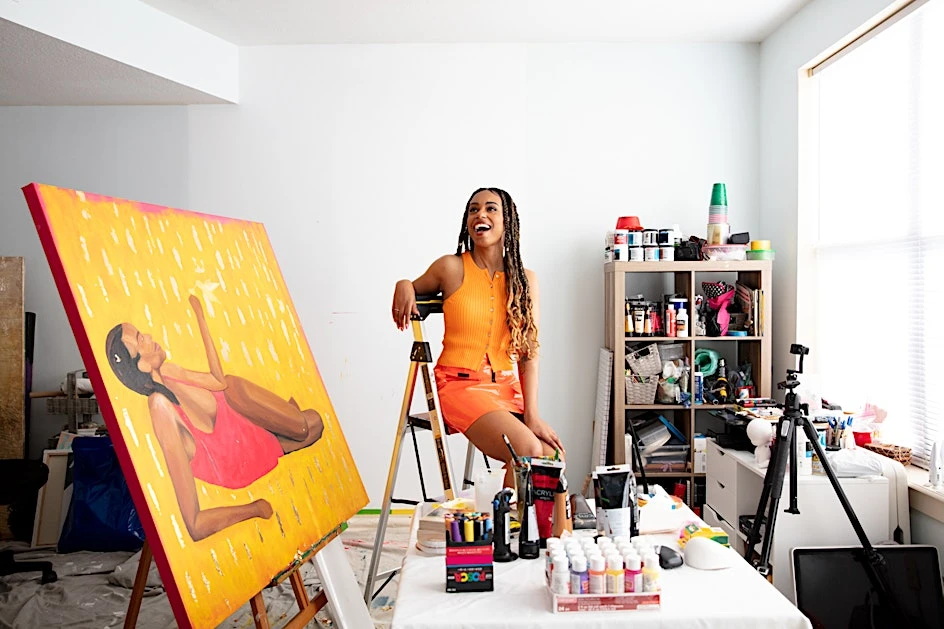 Happy artist poses in her studio next to a vibrant yellow and pink painting of a woman, surrounded by paint supplies and equipment.