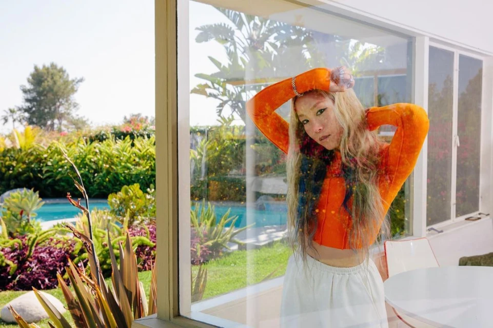 A young woman with blonde hair poses inside by a window, wearing an orange top and white skirt, with a garden and pool visible outside.
