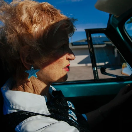 An elderly woman with red lipstick and star earrings drives a teal vintage car on a sunny day, wearing a white and black patterned jacket.