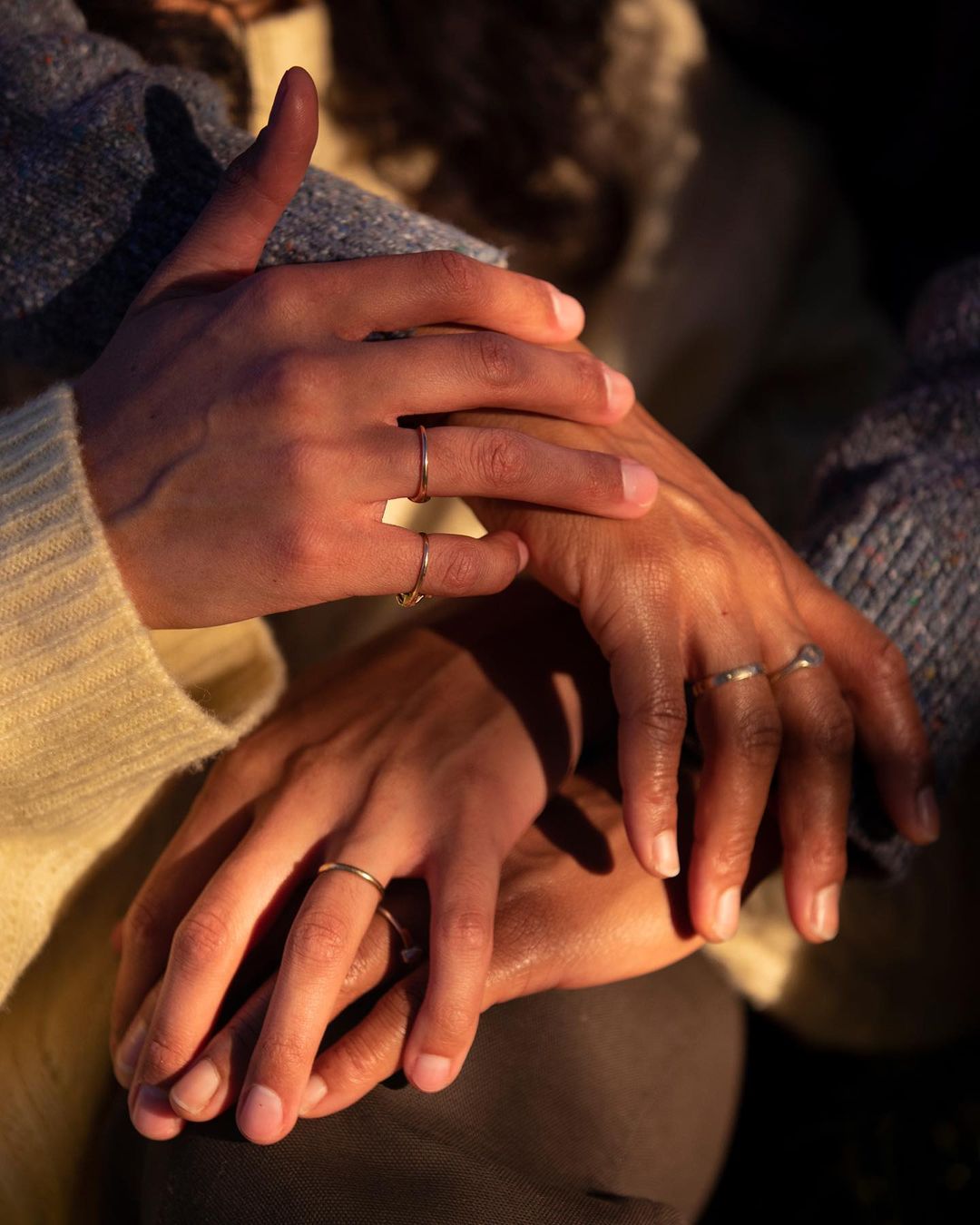 Close-up of two people holding hands with rings, warm-toned sunlight, neutral-color sweater, showing the connection and jewellery detail.