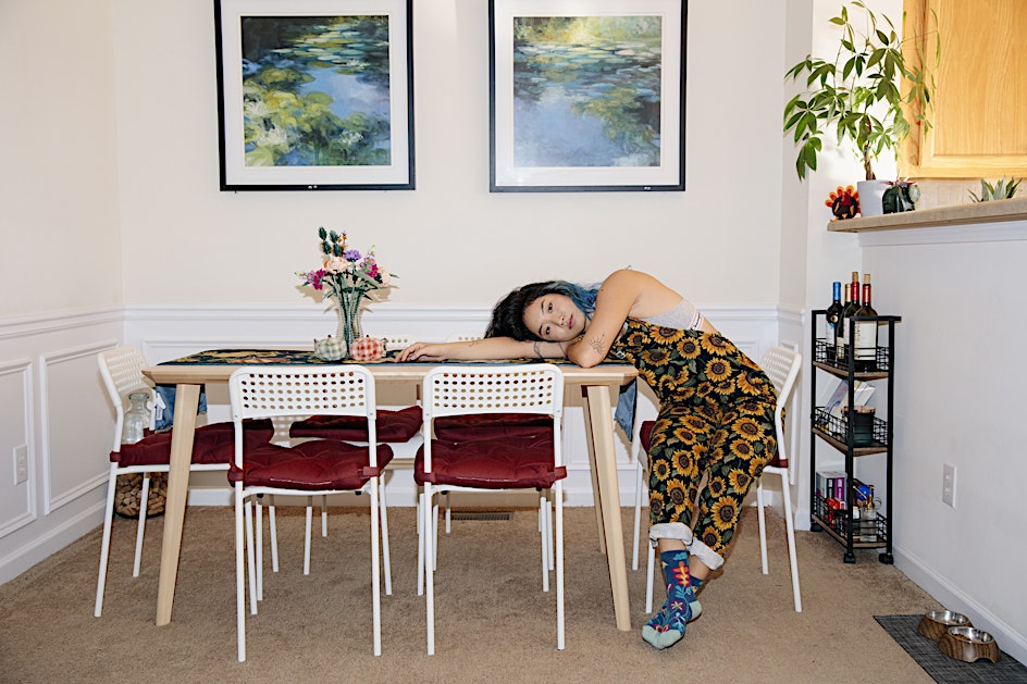 Woman with blue hair lies on dining table wearing sunflower overalls in indoor setting with artwork and potted plants, medium shot.