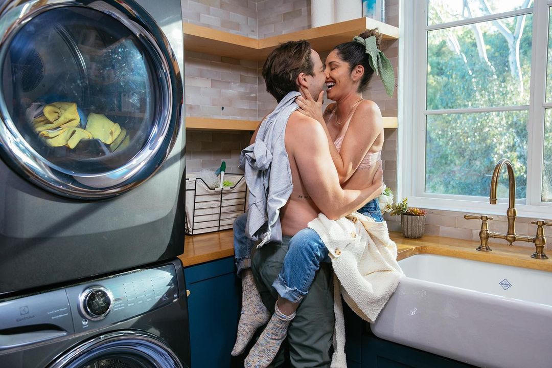 Affectionate couple in a sunlit laundry room embracing near washer/dryer, white sink, and open window showing trees outside.