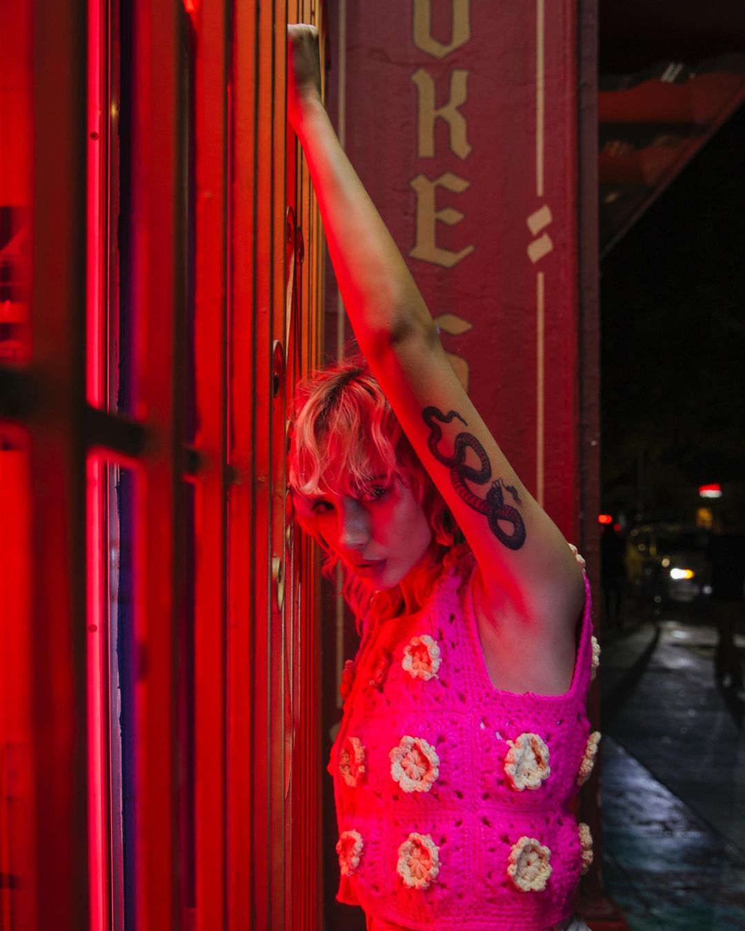 Stylish woman in a pink floral-accented crochet top poses near a red door frame at night in an urban setting with visible tattoo art.