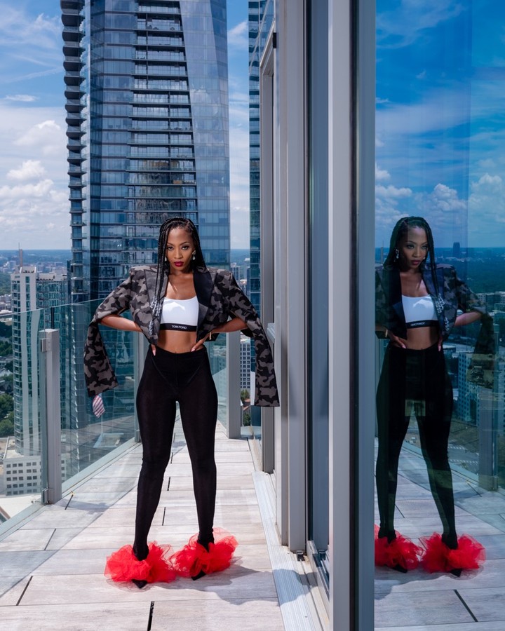 A black female model poses on a balcony with a tall skyscraper in the background, wearing a patterned blazer, leggings, and red fluffy slippers.