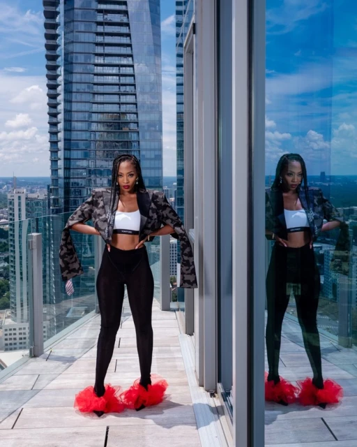 A black female model poses on a balcony with a tall skyscraper in the background, wearing a patterned blazer, leggings, and red fluffy slippers.