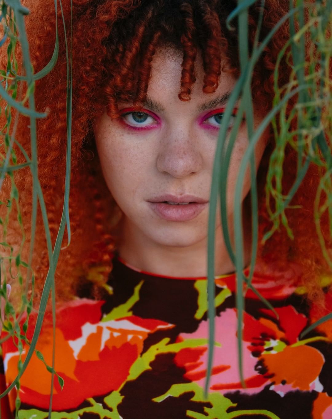 Close-up portrait of a woman with curly red hair and pink eye shadow, framed by green foliage, wearing a floral-patterned top.