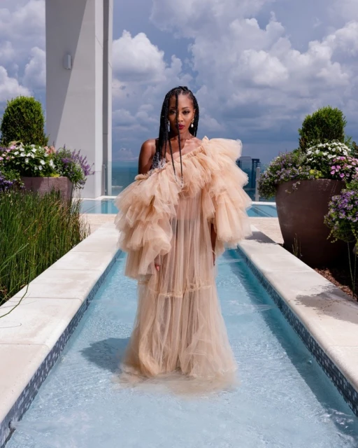 An adult female stands in shallow water, wearing a beige, ruffled gown, on a rooftop terrace with greenery and a city view.