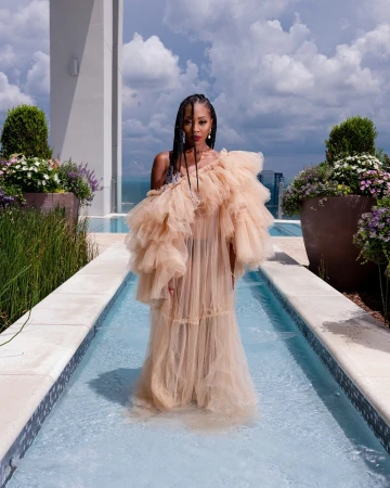 An adult female stands in shallow water, wearing a beige, ruffled gown, on a rooftop terrace with greenery and a city view.