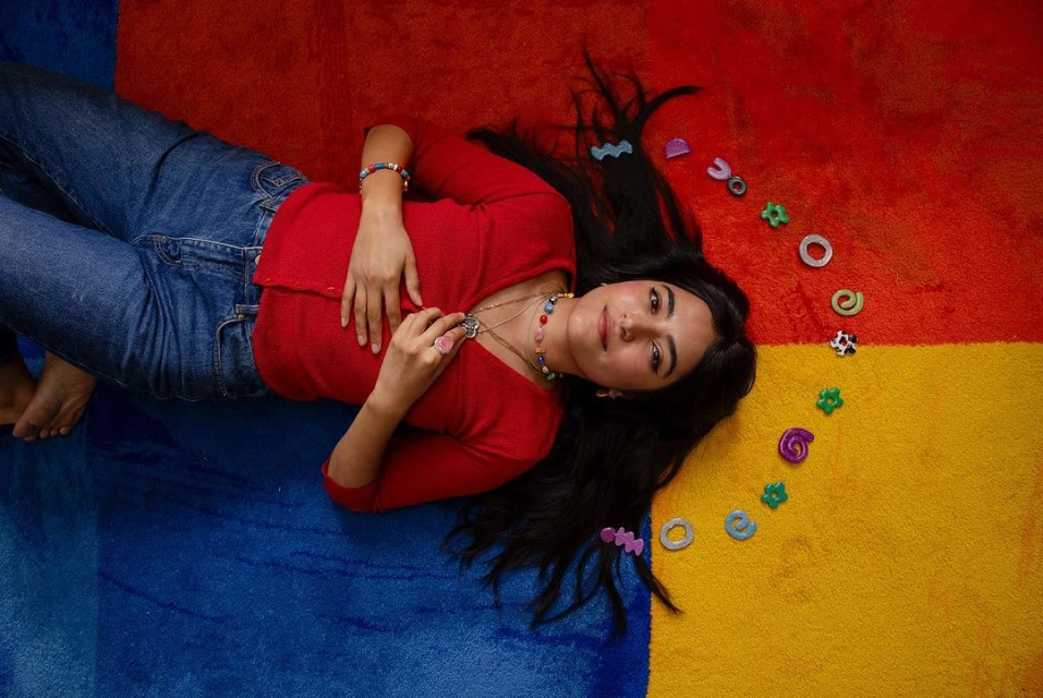 Young woman lying on a colorful rug surrounded by jewelry pieces, wearing a red top and jeans in an eye-level, medium shot.