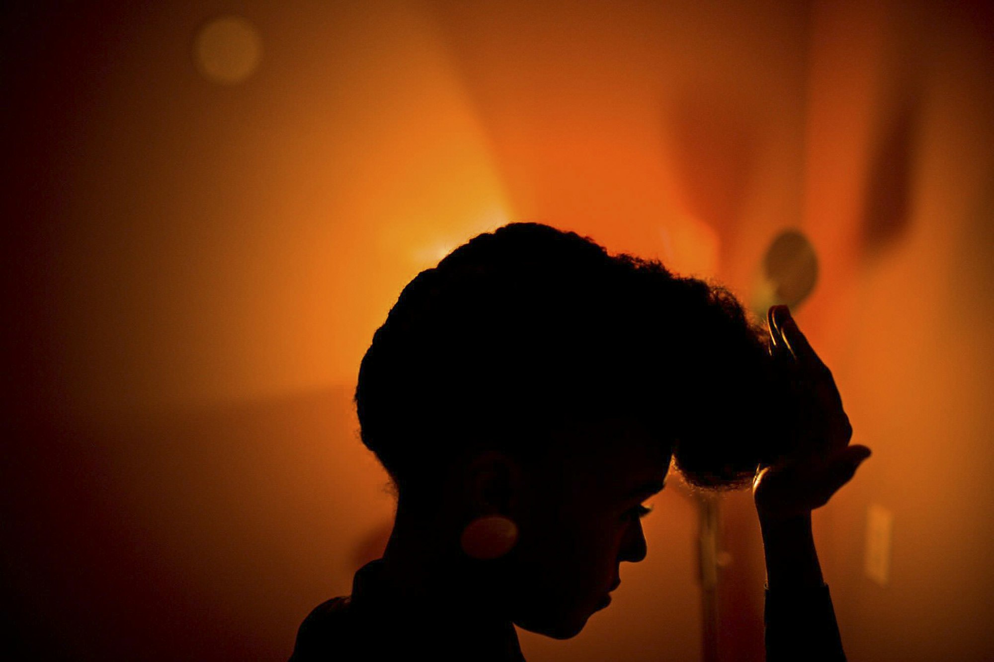 Silhouette of an adult female with afro hairstyle posing indoors under warm lighting, appearing contemplative in the dark environment.