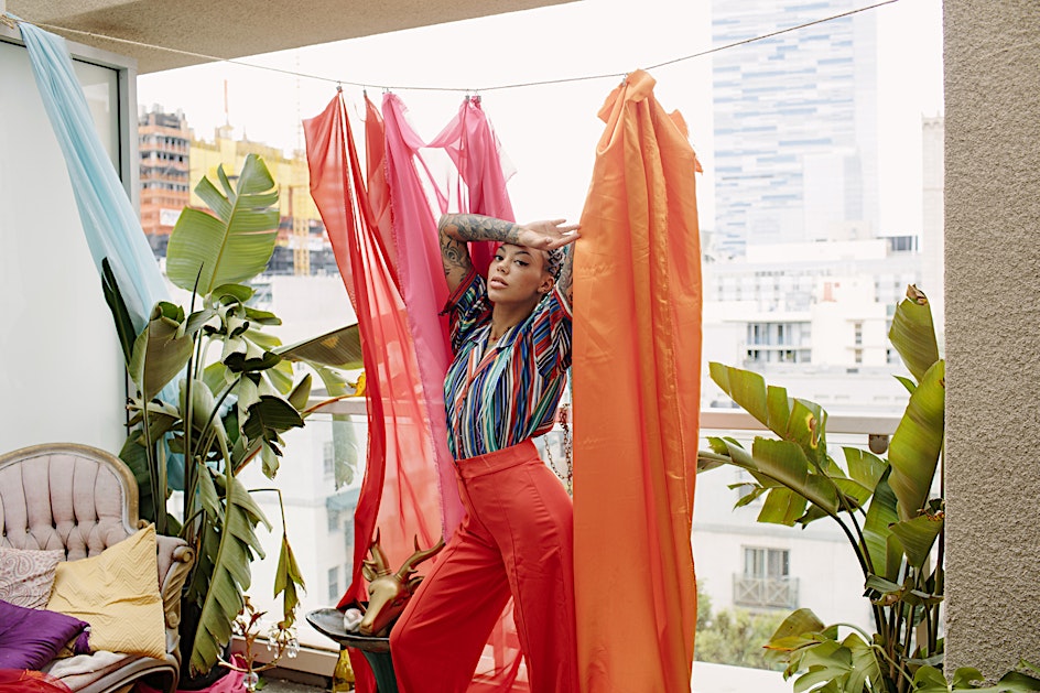 Adult female posing in orange pants and striped shirt on a balcony with tropical plants and sheer fabric with a city view.