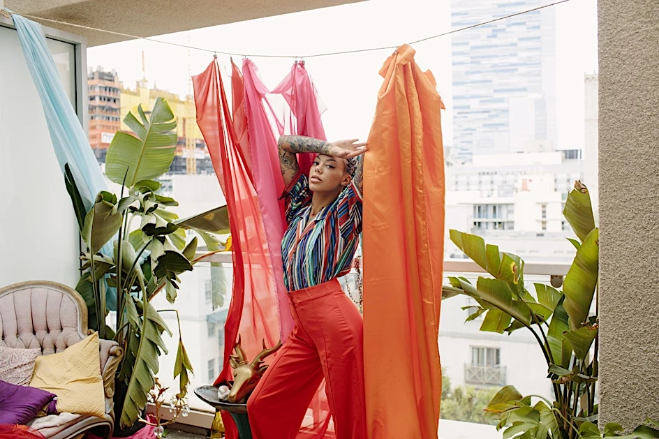Adult female posing in orange pants and striped shirt on a balcony with tropical plants and sheer fabric with a city view.