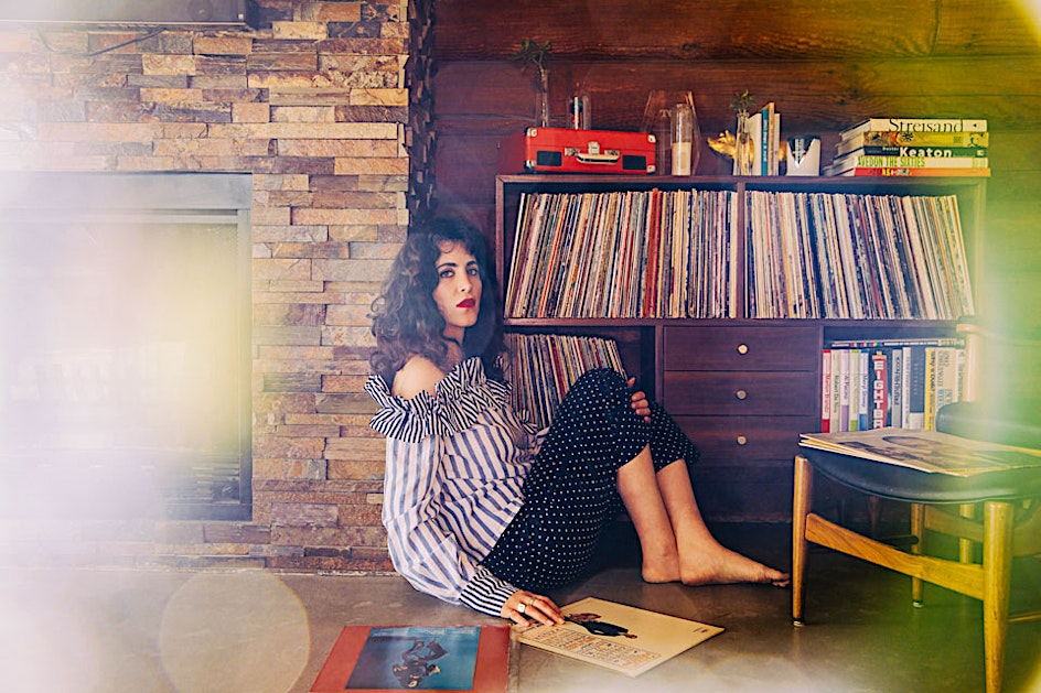 A woman with curly hair sits on the floor next to a brick wall and a record collection. Records are also on the floor beside her.