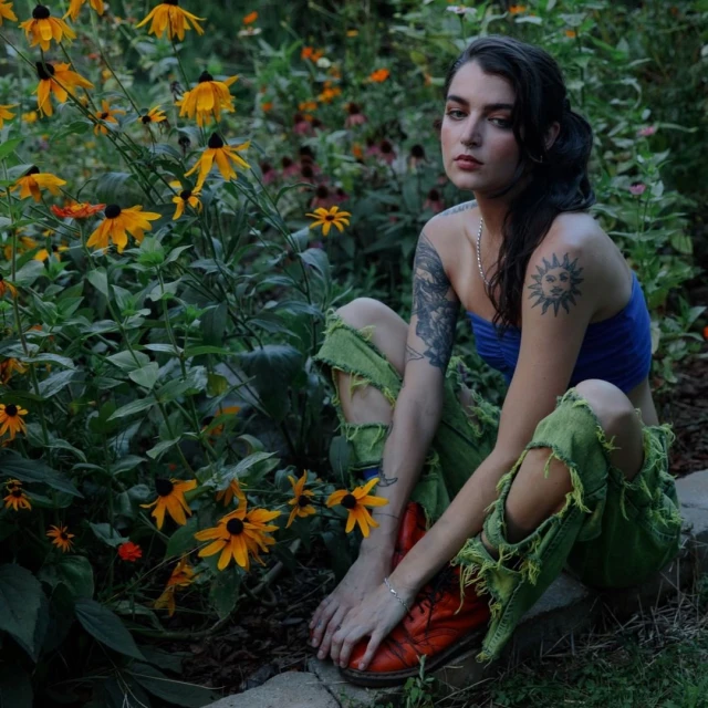 Adult female with shoulder tattoos poses in a garden of Black-Eyed Susans wearing ripped jeans, and orange boots in a medium outdoor shot.