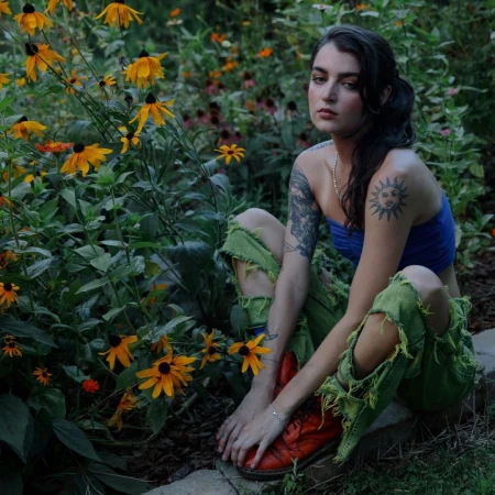 Adult female with shoulder tattoos poses in a garden of Black-Eyed Susans wearing ripped jeans, and orange boots in a medium outdoor shot.