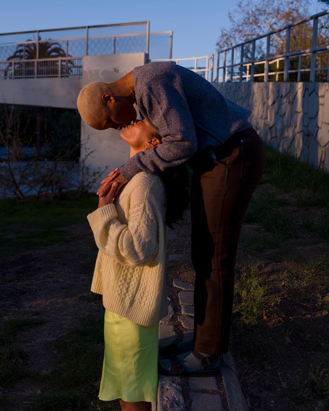 A couple embraces and kisses outdoors, dressed in casual sweaters and skirt, in front of a garden setting with trees and clear sky.