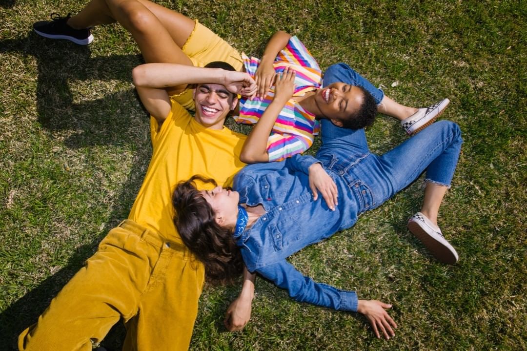 Three young friends lying on a green grass lawn laughing together in an overhead shot captured by Brinson+banks