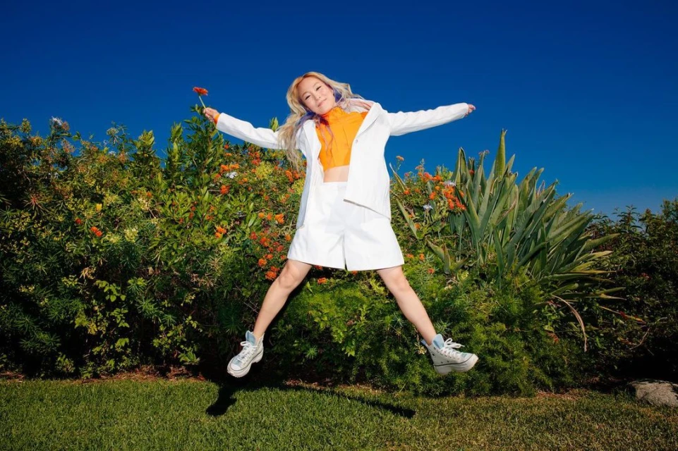 A blonde woman in a white shorts suit jumps in a grassy field with foliage and blue sky, holding a small orange flower.