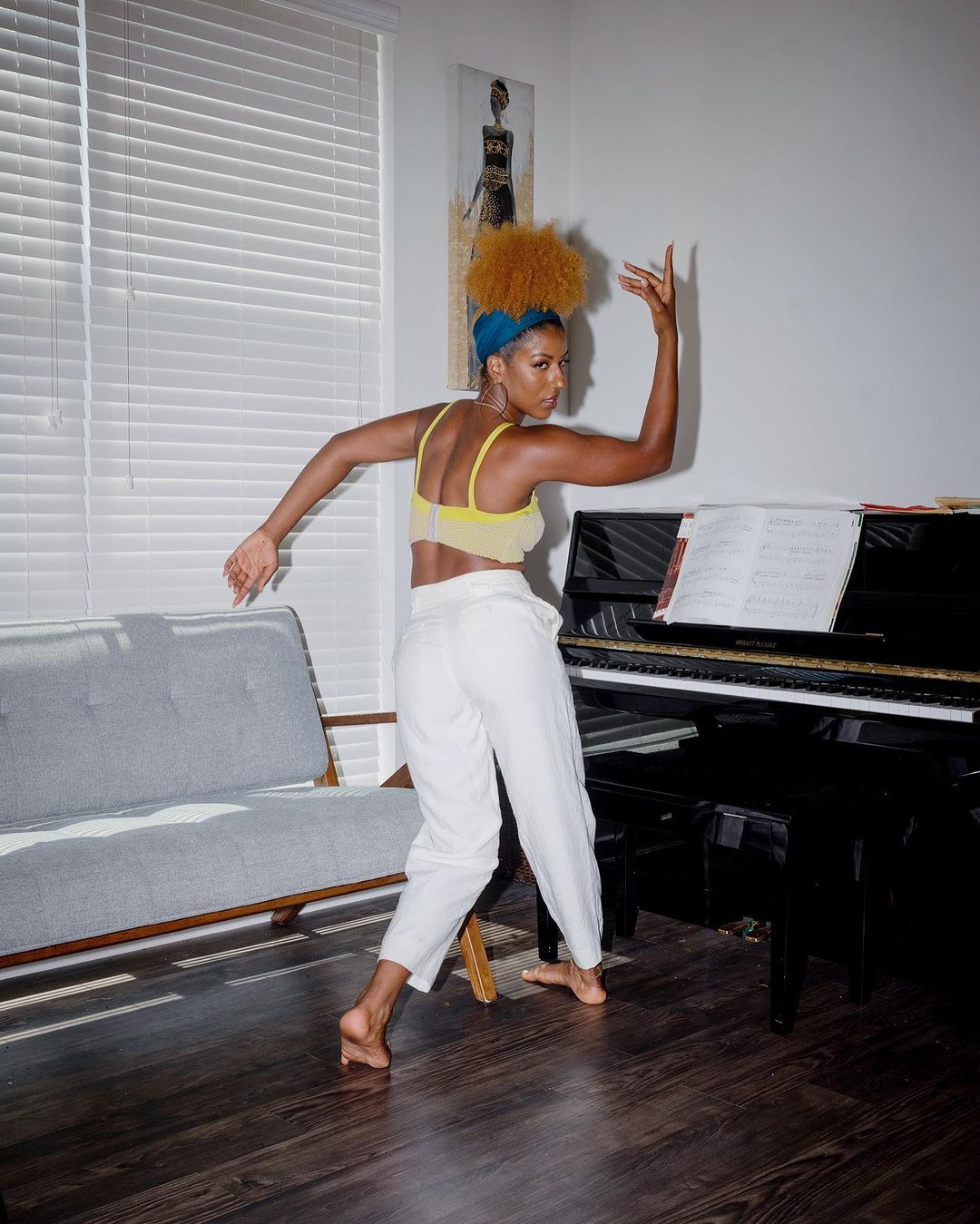 An African-descent woman with an afro dances barefoot in a yellow top and white pants, with a gray couch and piano visible in the background.