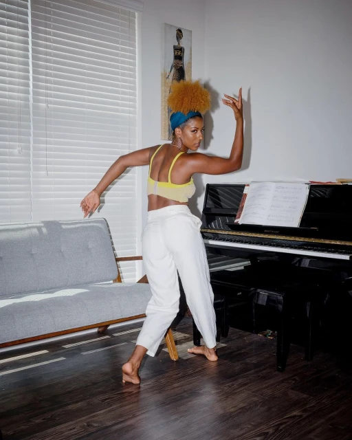 An African-descent woman with an afro dances barefoot in a yellow top and white pants, with a gray couch and piano visible in the background.