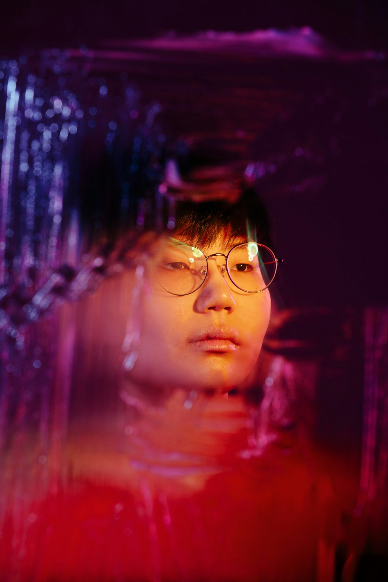 Portrait of a person with short dark hair and glasses, lit in red and purple tones, against a blurry background.