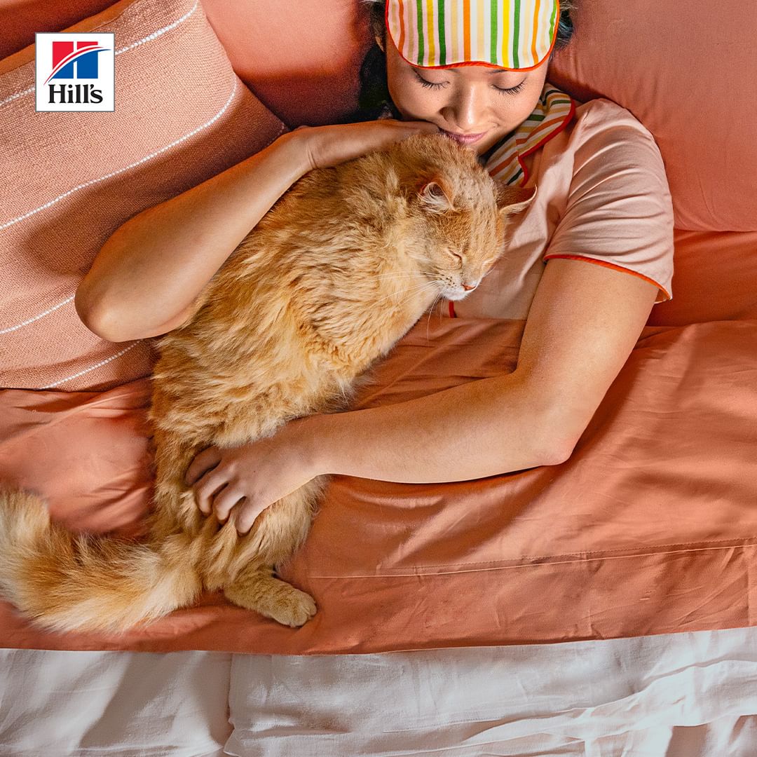 A woman wearing a sleep mask is hugging an orange tabby cat while laying in bed with peach-colored sheets and pillows in soft light.
