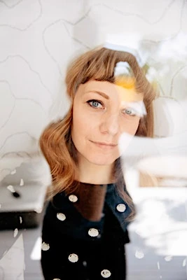 Portrait of a fair-skinned woman with wavy auburn hair looking through a window with a white wall featuring grey line art behind her.