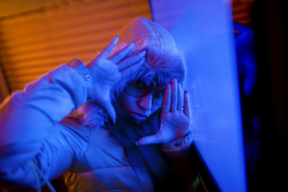 Portrait of a person in a fur-lined hooded jacket framing their face with their hands, lit with blue and orange light in a dark indoor space.