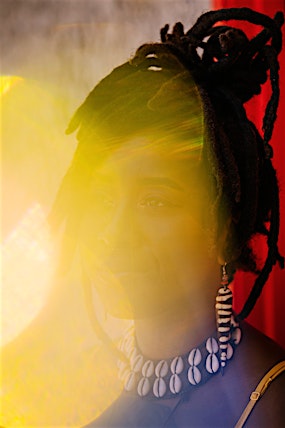Portrait of a woman with dreadlocks wearing a cowrie shell necklace and long earrings, partially obscured by bright yellow light leak.