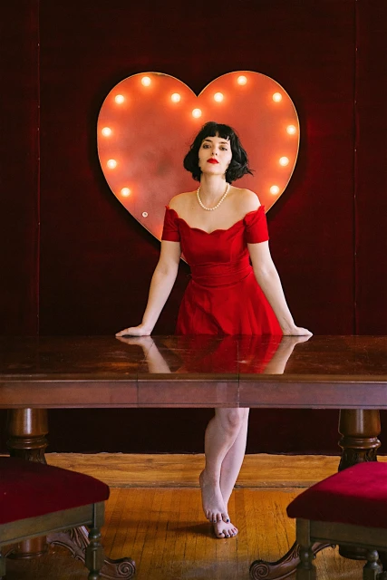 Elegant woman in a red dress poses next to a wooden table and an illuminated heart-shaped sign against a dark red background.