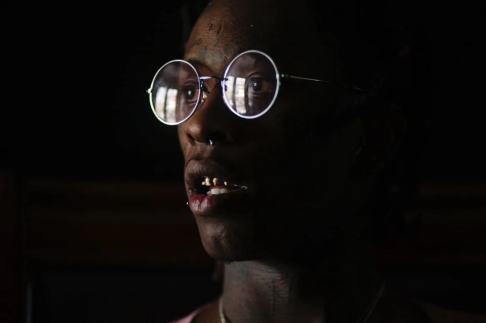 Close-up shot of a man wearing round glasses and gold teeth, set against a stark black background, emphasizing his facial features.