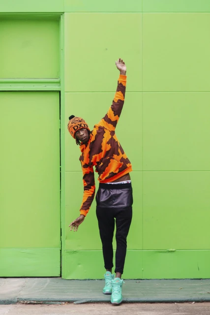 A person in an orange and brown patterned hooded sweatshirt and hat poses against a bright green wall outdoors in a full-body shot.