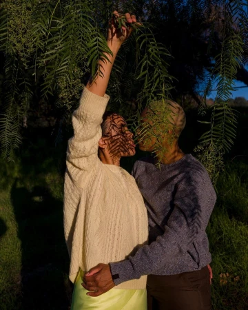 Romantic outdoor image of a couple embracing under leafy branches. One figure wears a light sweater and skirt; the other, a dark sweater.